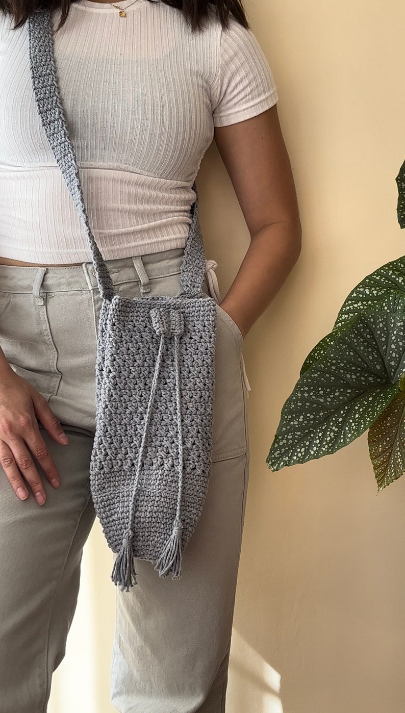 Peut inclure: Un sac &agrave; bandouli&egrave;re en crochet gris avec une fermeture &agrave; cordon et des accents de pompons.