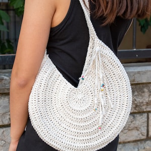 May include: Cream-colored, crocheted shoulder bag with a circular design. The bag features a long strap and a decorative fringe with colorful beads. The bag is worn over the shoulder, showcasing its handmade texture and bohemian style.