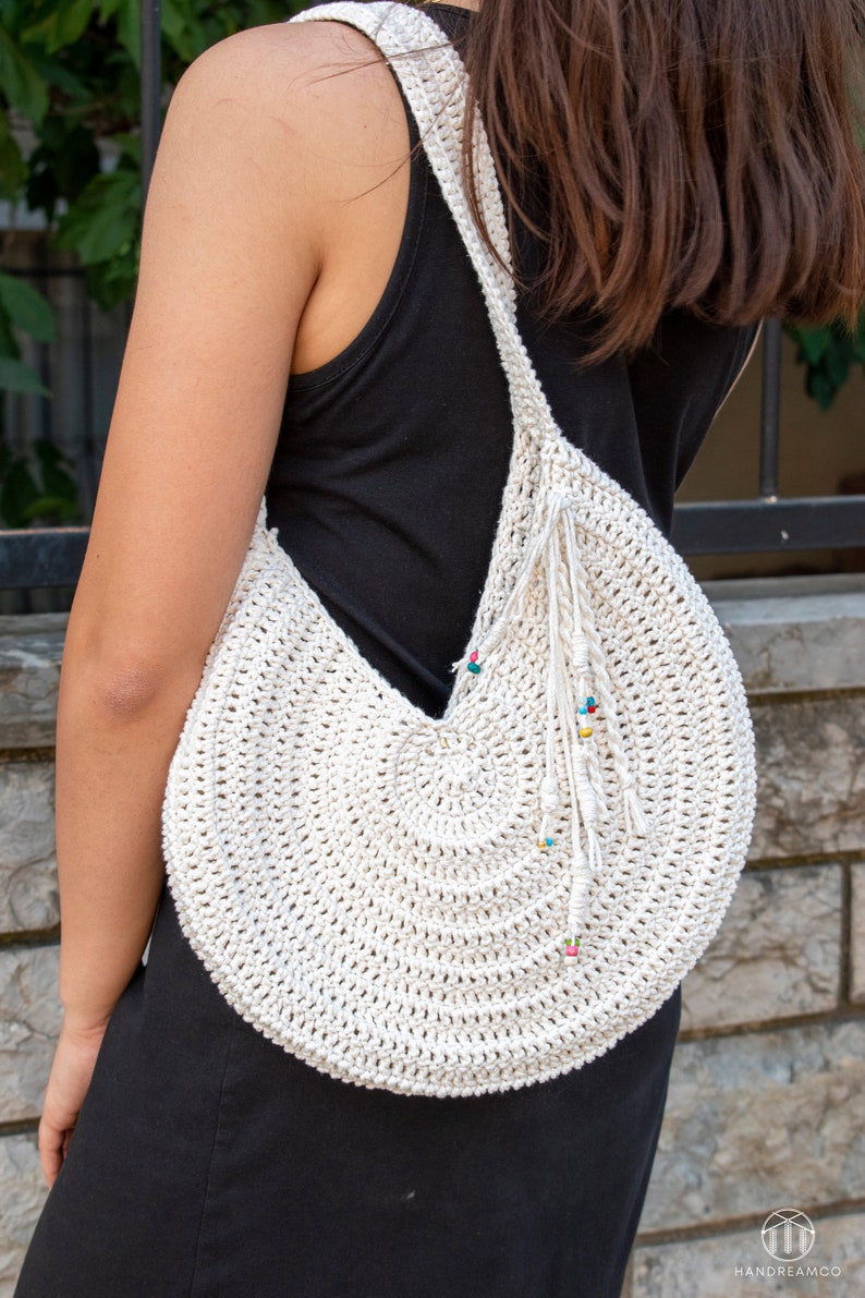 May include: Cream-colored, crocheted shoulder bag with a circular design. The bag features a long strap and a decorative fringe with colorful beads. The bag is worn over the shoulder, showcasing its handmade texture and bohemian style.