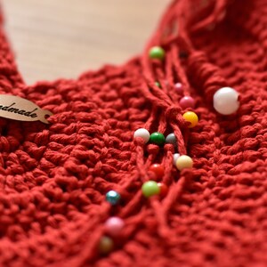 May include: Close-up of a handmade red crochet item with a tag that says "Handmade." The item features a textured stitch pattern and is adorned with strands of colorful beads in various sizes and colors. The overall theme is handcrafted textile art.