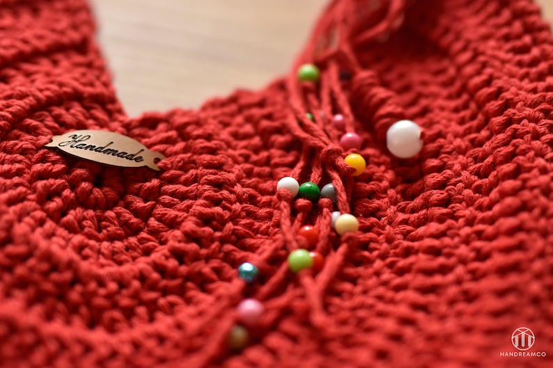 May include: Close-up of a handmade red crochet item with a tag that says "Handmade." The item features a textured stitch pattern and is adorned with strands of colorful beads in various sizes and colors. The overall theme is handcrafted textile art.