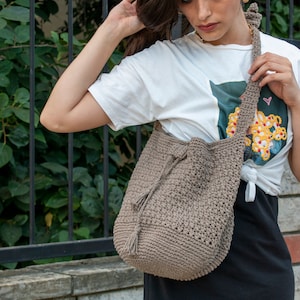 Peut inclure: Un sac cabas en crochet marron avec un pompon. Le sac est port&eacute; sur l'&eacute;paule d'une personne portant un t-shirt blanc &agrave; imprim&eacute; floral et une jupe noire.