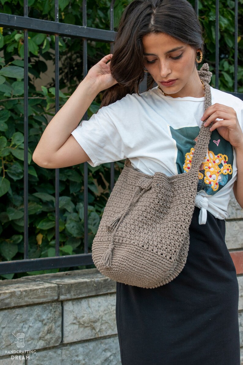 Peut inclure: Un sac cabas en crochet marron avec un pompon. Le sac est port&eacute; sur l'&eacute;paule d'une personne portant un t-shirt blanc &agrave; imprim&eacute; floral et une jupe noire.