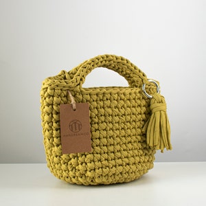 Pu&ograve; includere: Una piccola borsa a mano in crochet gialla con un'etichetta in pelle marrone che dice "HANDREAMCO". La borsa ha un manico superiore e un pon pon.