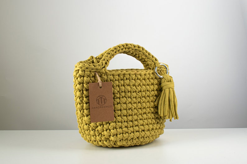 Pu&ograve; includere: Una piccola borsa a mano in crochet gialla con un'etichetta in pelle marrone che dice "HANDREAMCO". La borsa ha un manico superiore e un pon pon.