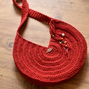 May include: A handmade red crochet shoulder bag with a long strap. The bag features a circular design and is adorned with a small wooden tag and colorful beads. The bag is made of a thick, textured yarn.
