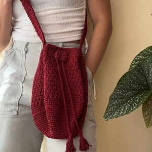 Peut inclure: Un sac en crochet de couleur bordeaux avec une fermeture &agrave; cordon et des accents de pompons. Le sac est port&eacute; en bandouli&egrave;re.