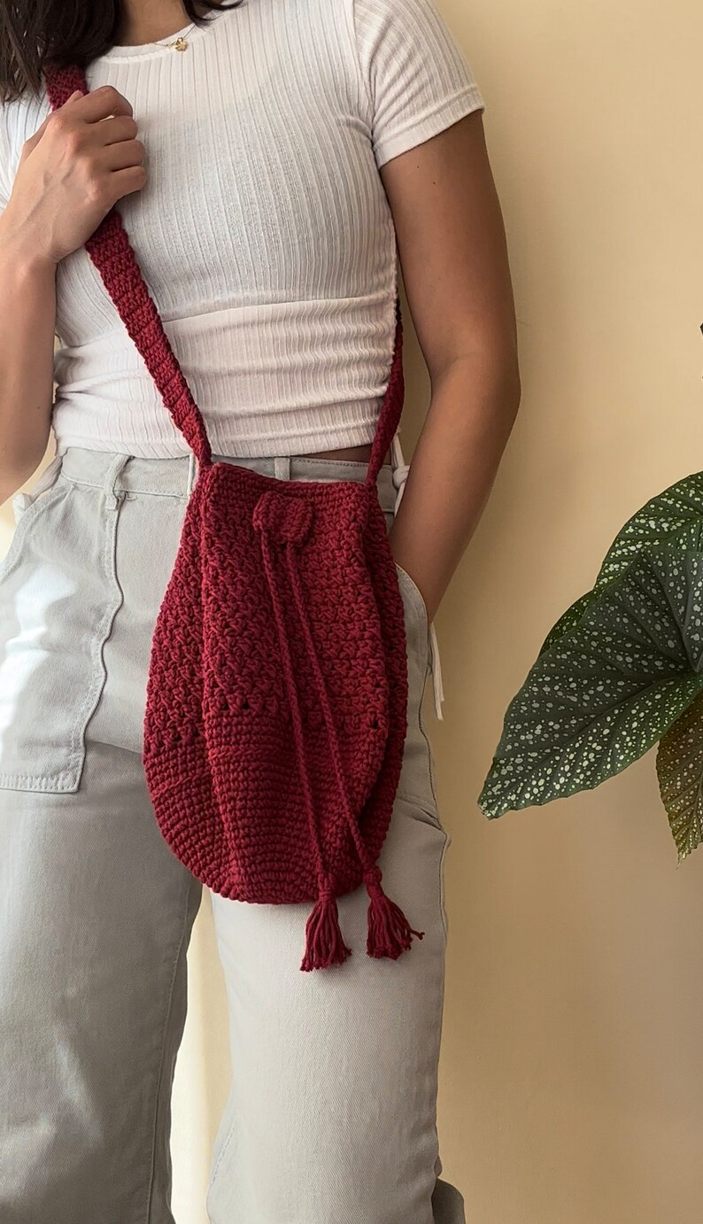 Peut inclure: Un sac en crochet de couleur bordeaux avec une fermeture &agrave; cordon et des accents de pompons. Le sac est port&eacute; en bandouli&egrave;re.