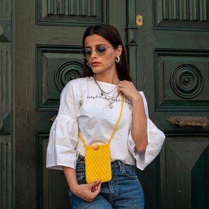 Puede incluir: Una mujer con una camiseta blanca con el texto "Good girls go to heaven, bad girls go everywhere" y pantalones vaqueros azules sostiene un bolso de hombro de crochet amarillo.