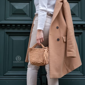 Pu&ograve; includere: Una donna indossa un maglione bianco, pantaloni beige e un cappotto color cammello. Tiene una borsa di uncinetto marrone con un fiocco.