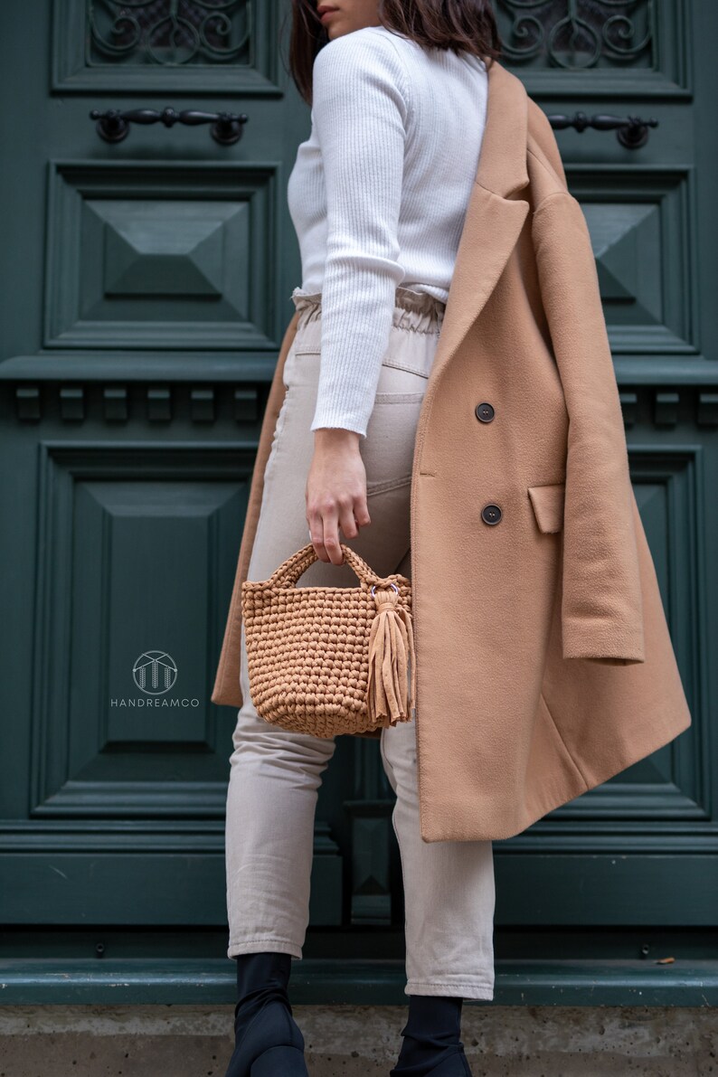 Pu&ograve; includere: Una donna indossa un maglione bianco, pantaloni beige e un cappotto color cammello. Tiene una borsa di uncinetto marrone con un fiocco.