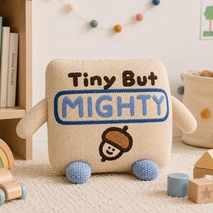 May include: A square, beige knit plush toy with two arms and two blue feet. The front features the text "Tiny But MIGHTY" in dark brown and light blue, with a small, smiling brown acorn graphic positioned below the text on the knit surface.