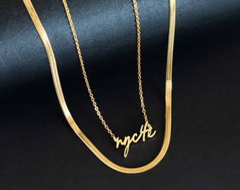 Puede incluir: Dos collares de oro, uno con una cadena delicada y un colgante que dice "nycth", y el otro con una cadena más gruesa y texturizada.