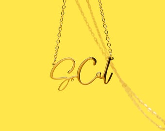 Puede incluir: Un collar dorado con las letras cursivas "S. Cal" suspendidas sobre un fondo amarillo brillante. La cadena es delicada y las letras están conectadas. El collar está centrado en el marco.