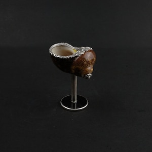 Puede incluir: Una concha de mar marrón con un borde plateado está montada en un soporte plateado. La concha de mar se utiliza como una taza decorativa.