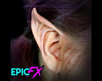 May include: Close-up of a pointed, elf-like ear prosthetic made of silicone. The ear is pale pink and has a natural texture. The ear is attached to a person's head, with purple hair visible.