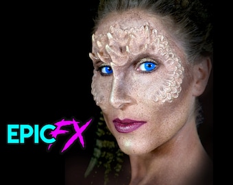 May include: A woman with blue contact lenses and white makeup on her face. The makeup is designed to look like scales. The woman is looking at the camera. The text "EPIC FX" is in the lower left corner of the image.
