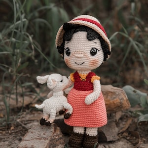 以下が含まれることがあります： 赤い白のドレスと茶色の帽子をかぶった、かぎ針編みのドールが、白い羊毛のかぎ針編みの羊を抱いています。ドールと羊は岩の上に立っています。