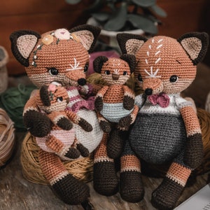 May include: A family of crocheted fox dolls in shades of brown, tan, and black. The dolls have detailed features, including embroidered faces and decorative accents. The largest fox wears a bow tie, while others have floral headbands. The dolls are arranged on a wooden surface.
