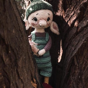 Könnte beinhalten: Eine gehäkelte Elfenpuppe mit grünem Hut, grüner und lila Latzhose und roten Schuhen. Die Puppe steht in einem Baumstamm.