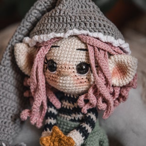 Puede incluir: Una muñeca elfo de crochet con pelo rosa, un gorro gris y un atuendo a rayas verde y negro. El elfo sostiene una estrella dorada.