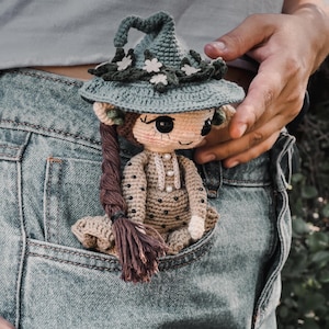 Puede incluir: Una muñeca de crochet con un sombrero de bruja verde con flores blancas y un vestido marrón con lunares blancos. La muñeca está en un bolsillo de mezclilla azul.