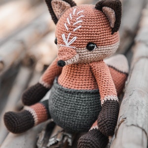 May include: A crocheted fox toy in brown, tan, and grey. The fox has a detailed face with white embroidered details and black eyes. It is wearing a grey nappy and has dark brown paws and ears. The doll is sitting on a wooden surface.