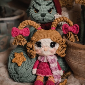 Könnte beinhalten: Eine gehäkelte Puppe mit braunen Haaren, einem rosa Kleid und einem rosa-weißen Schal. Die Puppe sitzt neben einer gehäkelten Grinch-Puppe mit einem roten Hut.