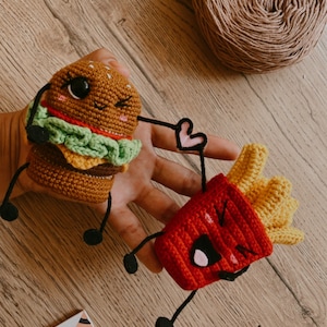 Puede incluir: Dos juguetes de comida de ganchillo, una hamburguesa y una caja de patatas fritas, con caras sonrientes y brazos y piernas de hilo negro. Los juguetes están sobre una superficie de madera.