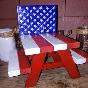 May include: A miniature red, white, and blue picnic table with a wooden backboard painted with the American flag. The table is made of wood and has a white and red striped design.
