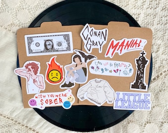 May include: A collection of colorful stickers featuring various designs, including a cartoon face, a person with a microphone, and the text "Conan Gray" and "Little League".