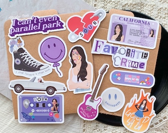 May include: A set of stickers featuring various designs, including a purple heart with the word "SOUR" on it, a purple smiley face balloon, a purple cassette tape with the word "SOUR" on it, a purple and white guitar with the words "It's Sour out here" on it, a purple and white band-aid with the words "hope ur ok" on it, a purple and white license plate with the words "Favorite Crime" on it, a purple and white sticker with the words "Sociopath" on it, a purple and white sticker with the words "I can't even parallel park" on it, a purple and white sticker with the words "God 4 U" on it, and a purple and white sticker with a cartoon image of a person with the words "Olivia Rodrigo" on it.
