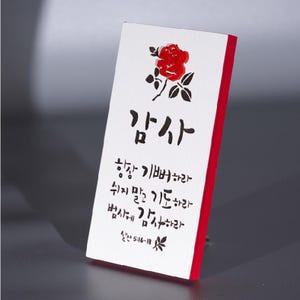 May include: A white rectangular plaque with red edges, featuring Korean calligraphy and a red rose illustration. The text includes the word "Thanks" and scriptural verses. A decorative item.
