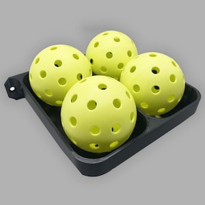 May include: Four yellow pickleball balls with holes in them are in a black plastic holder.