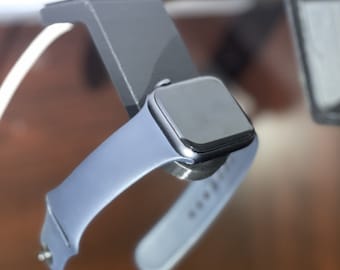 Op de afbeelding: Een zilveren Apple Watch met een lichtblauw sportbandje. Het horloge staat op een oplaadstandaard.