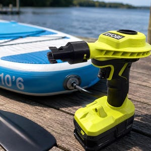 Pode incluir: Um inflador e deflator sem fio Ryobi amarelo neon e preto com um bocal preto, posicionado ao lado de uma prancha de stand up paddle inflável azul e branca com a etiqueta 10'6 e um remo preto em uma superfície de madeira.