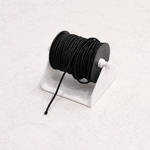 May include: A spool of black elastic cord sits on a white plastic holder. The cord is tightly wound around a black spool. A length of the cord extends from the spool, suggesting it is ready for use. The holder is a simple, angled design.