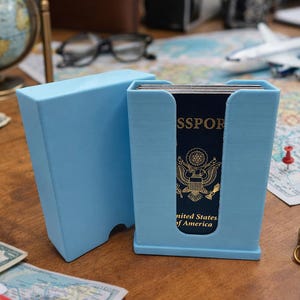 May include: A light blue passport holder with a matching lid, holding a United States passport. The holder is on a wooden surface with a globe, map, compass, and toy airplane in the background. The passport is visible.