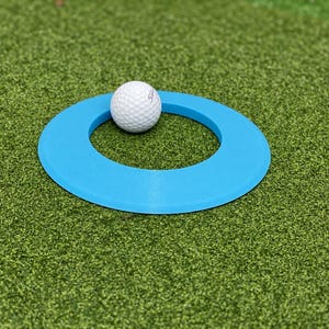 May include: A white dimpled golf ball resting inside a bright blue circular ring. The golf ball features the brand name Titleist printed in small black text on its surface. The blue ring has a flat, smooth finish and a wide, circular opening.