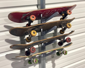 Könnte beinhalten: Eine Wandhalterung für Skateboards mit vier Skateboards. Die Skateboards sind in verschiedenen Farben, darunter Rot, Braun und Grün, mit sichtbaren Gebrauchsspuren. Die Halterung ist aus grauem Metall und an einer weißen Wand befestigt.