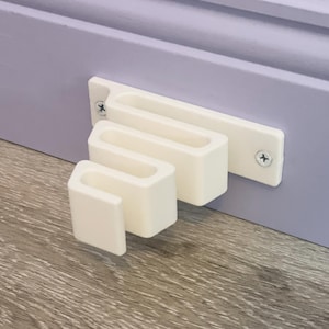May include: White plastic wall-mounted organizer with three slots of different sizes. The organizer is attached to a light purple wall with screws.