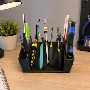 May include: A black plastic desk organiser holding various tools including pliers, a compass, a screwdriver with a blue and black handle, a green marker, and a metal ruler. The organiser has multiple compartments for upright storage of items.
