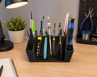 Può includere: Un organizer da scrivania in plastica nera contenente vari strumenti, tra cui pinze, un compasso, un cacciavite con manico blu e nero, un pennarello verde e un righello in metallo. L'organizer è dotato di scomparti per riporre gli oggetti.