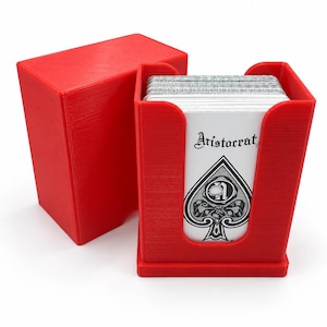 Könnte beinhalten: Ein leuchtend roter Spielkartenhalter aus Kunststoff mit passendem abnehmbarem Deckel. Der Halter enthält ein Kartenspiel mit weißen Karten, auf deren Vorderseite das Wort Aristocrat und ein schwarzes Pik-Symbol mit einer Schleife abgebildet sind.