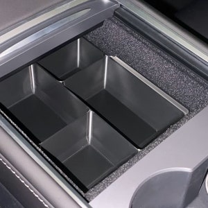 May include: A black plastic organizer with four compartments fits inside a car's center console. The organizer is designed to help keep the console tidy and organized.