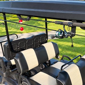 May include: A black golf cart featuring a roof-mounted fishing rod rack holding two fishing rods with reels and red and white bobbers. The cart has two rows of seats upholstered in alternating black and beige panels with white piping trim.