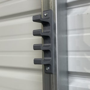 以下が含まれることがあります： 金属製の柱に2本のネジで取り付けられた、4つのスロットを持つ灰色のプラスチック製の壁掛け式オーガナイザー。工具やその他のアイテムを収納できます。