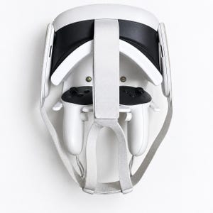 May include: A white virtual reality headset with a black face interface and a light gray fabric head strap, mounted on a wall-mounted storage bracket that also holds two white handheld controllers with black button panels and joysticks.