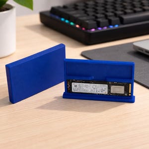May include: A blue SSD enclosure with an open case revealing the internal drive. The enclosure is made of a solid, smooth material. A black keyboard and a laptop are in the background.