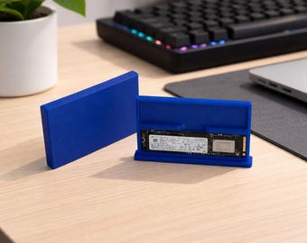 May include: A blue SSD enclosure with an open case revealing the internal drive. The enclosure is made of a solid, smooth material. A black keyboard and a laptop are in the background.